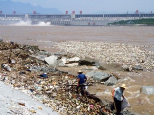 This image shows the Three Gorges Dam in the background with masses of waste floating and being washed up on the shore of the Yangtze River in the foreground.